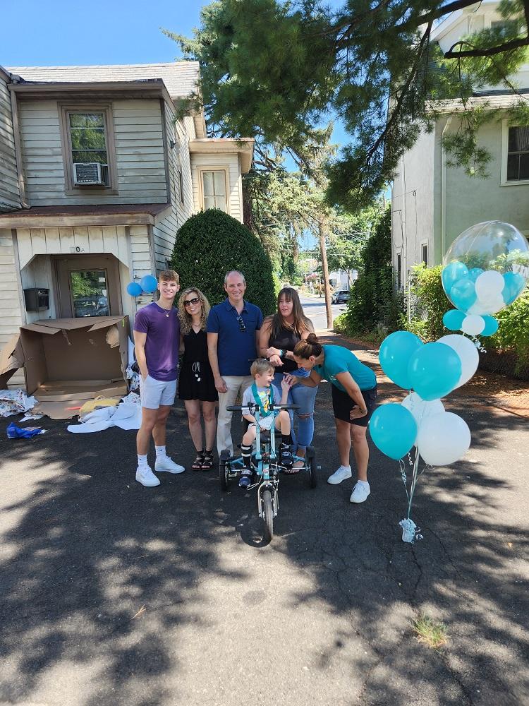 Help Hope Live Executive Director Kelly L Green reaches down to hold Vinny’s hand as the 4-year-old sits on his new Freedom Concepts teal adaptive bike. Vinny wears a t-shirt and shorts with a black leg brace on his lower right leg. Kelly is wearing a teal Help Hope Live t-shirt. She has light skin with her brown hair is pulled back into a bun. Behind Vinny are Help Hope Live supporters and Vinny’s mom. Beside them are teal and white balloons. They are outdoors on the blacktop in front of Vinny’s house with a large opened cardboard box behind them. A blue sky and trees are visible.