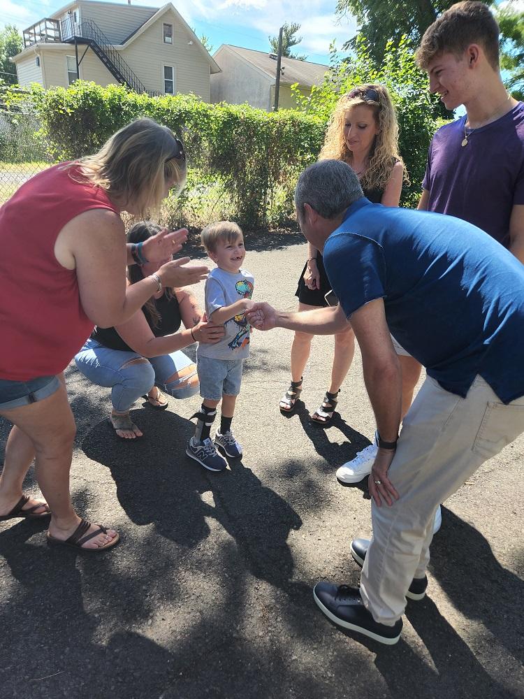 4-year-old Vinny gets a handshake from a Help Hope Live supporter and smiles. He is outside with his family wearing shorts and a t-shirt with a black leg brace on his lower right leg.
