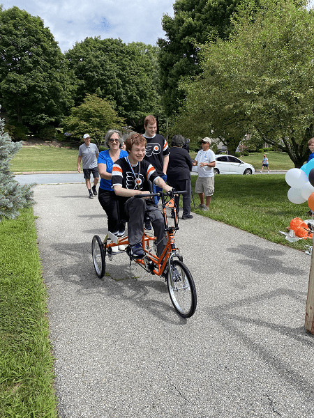 18-year-old Patrick grins as he rides his orange and black Freedom Concepts adaptive bike. He’s wearing a black and orange Flyers jersey and a replica gold medal. He’s being pushed by a loved one with gray hair and a blue shirt.