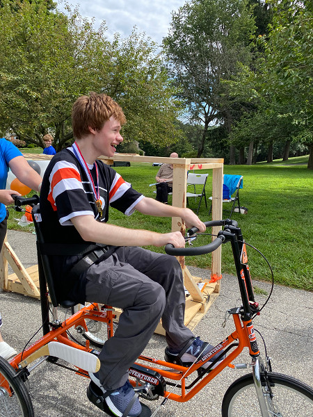 18-year-old Patrick grins as he rides his orange and black Freedom Concepts adaptive bike. He’s wearing a black and orange Flyers jersey and a replica gold medal. Patrick has red hair and light skin.