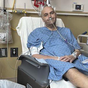 Liver transplant recipient Brent is in the hospital seated wearing a blue hospital gown with a shaved head and light skin. He is hooked up to several machines.