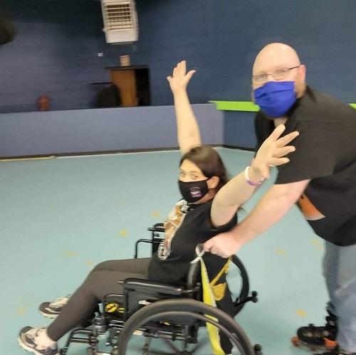 Kidney transplant recipient Jackie raises her arms gleefully while seated in a black manual wheelchair being pushed by a male presenting person. Jackie is wearing a black mask and her wheelchair pusher is wearing a blue mask.