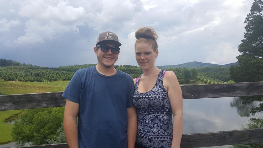 Transplant candidate Zachary Campbell is pictured with his wife in a beautiful outdoor setting. Zach wears a brown hat, black sunglasses, and a blue t-shirt. His wife wears a tank top with a white geometric pattern and has red hair up in a bun.