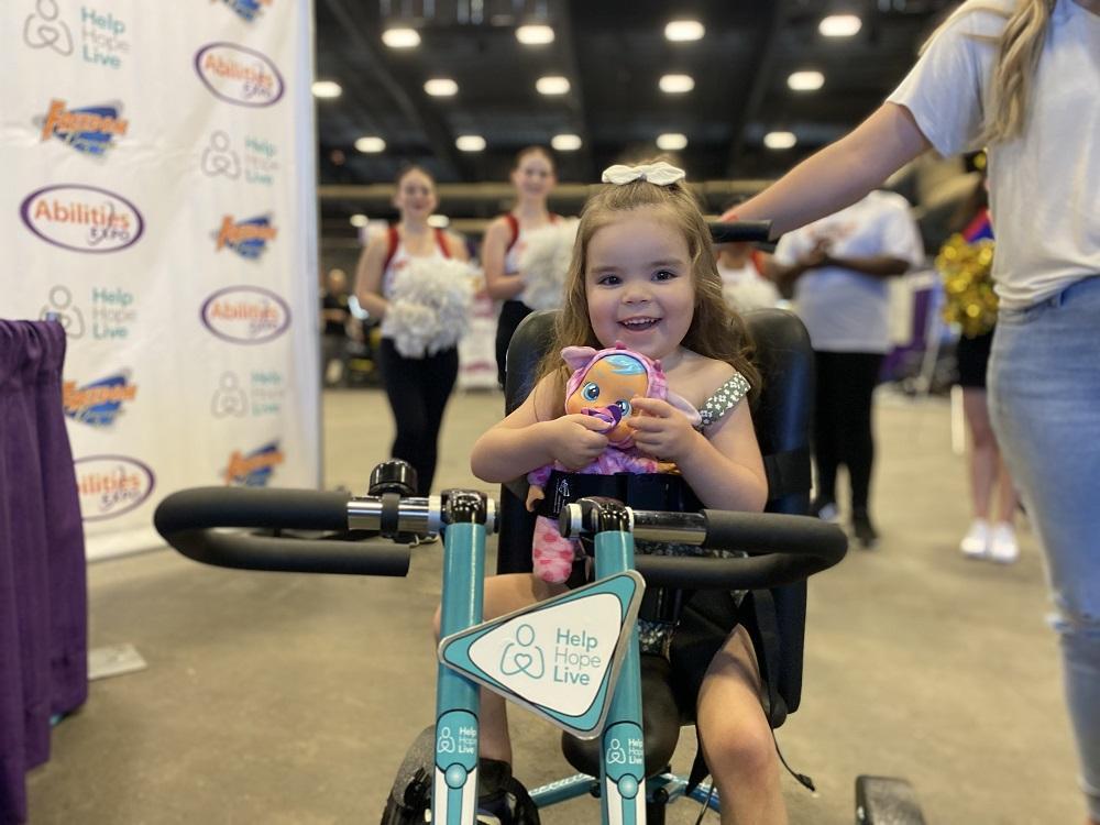 3-year-old Emberly smiles holding a doll as she sits on her new adaptive bike. She is at the Abilities Expo Phoenix.