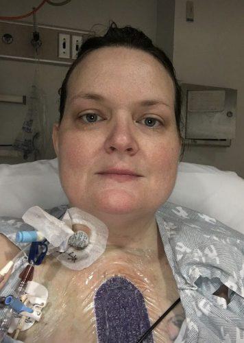 Heart transplant recipient Linda Jara in the hospital wearing a gown and hooked up to multiple devices. She has blue-gray eyes, light skin, and brown hair pulled back.