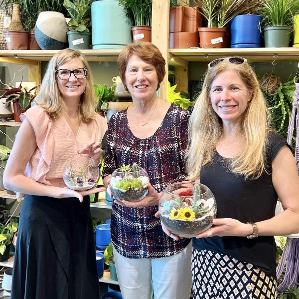 Double transplant recipient Amanda Ross is pictured with two women, all holding small circular glass planters with plants visible behind them. Amanda has shoulder length straight blonde hair, light skin, and black rimmed glasses.