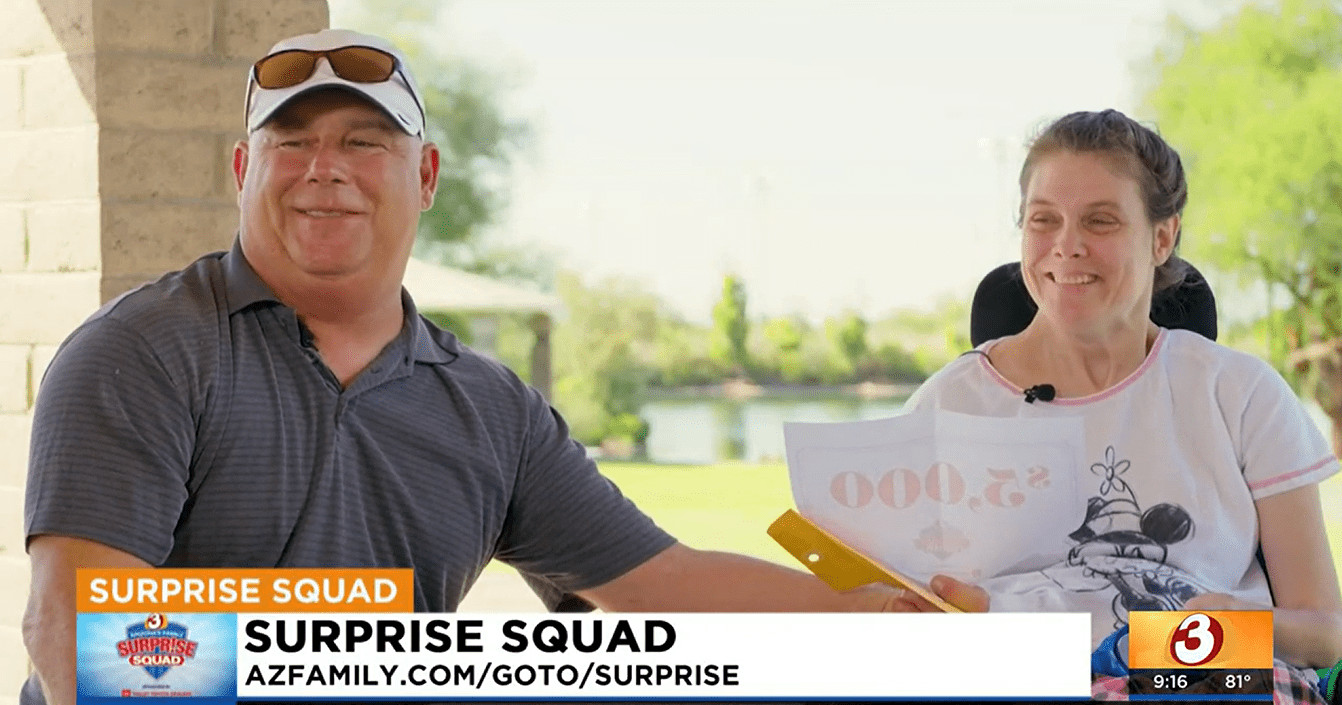 Help Hope Live spinal cord injury client Lori Vehmeier grins as she looks at a paper that reads $5,000 the amount of the donation to Help Hope Live in her honor to fully fund the mattress she needs. To her right is her uncle Verne who is also smiling. Lori sits in a black power chair with a park behind her. The ticker reads SURPRISE SQUAD from AZ Family channel 3.