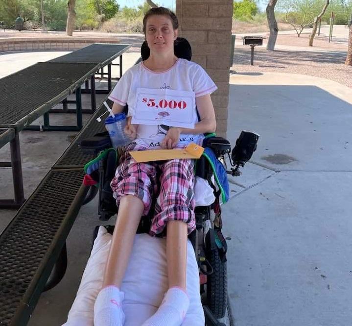 Help Hope Live spinal cord injury Lori Vehmeier smiles and holds a sign that reads $5,000 as she sits in her black power chair outdoors in a public park.