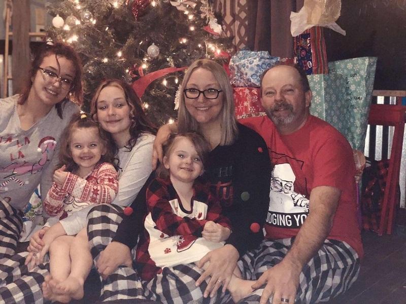 Pearl Brown is in a holiday scene with her family: her husband and four girls of various ages. They are in front of a Christmas tree. Pearl has light skin, blonde hair, and black glasses.