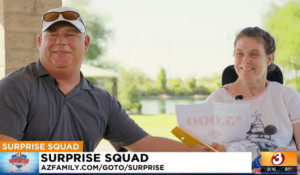 Help Hope Live spinal cord injury client Lori Vehmeier grins as she looks at a paper that reads $5,000 the amount of the donation to Help Hope Live in her honor to fully fund the mattress she needs. To her right is her uncle Verne who is also smiling. Lori sits in a black power chair with a park behind her. The ticker reads SURPRISE SQUAD from AZ Family channel 3.