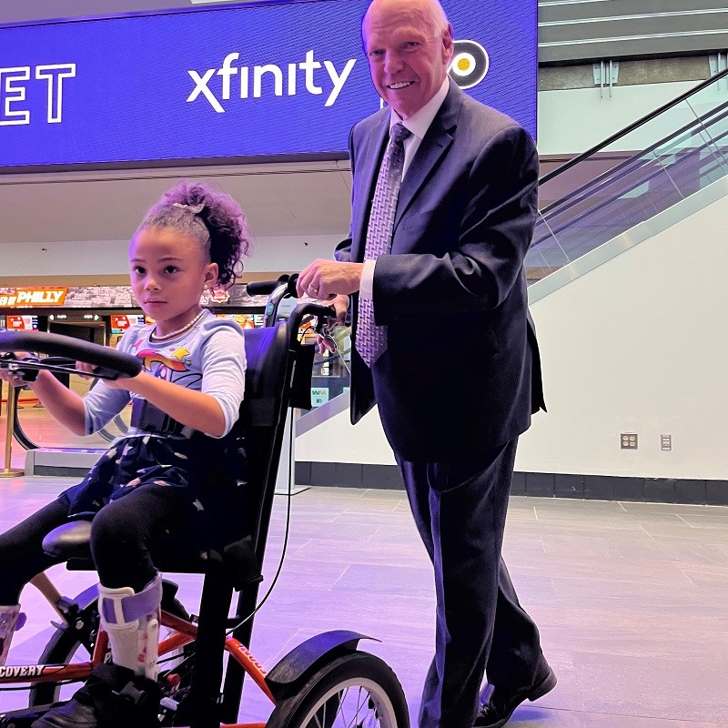 Bob Kelly of the Flyers Alumni smiles as he pushes 6-year-old Evangeline on her new adaptive bike.