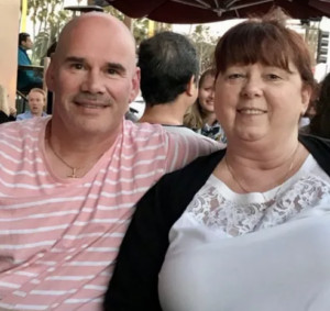 Bob Haines and his wife, Dawn, are seated at an outdoor establishment. Bob has light skin and a bald head and wears a light pink striped shirt. Dawn has light skin and red-brown hair pulled back and wears a white lace-trimmed shirt and a black sweater.