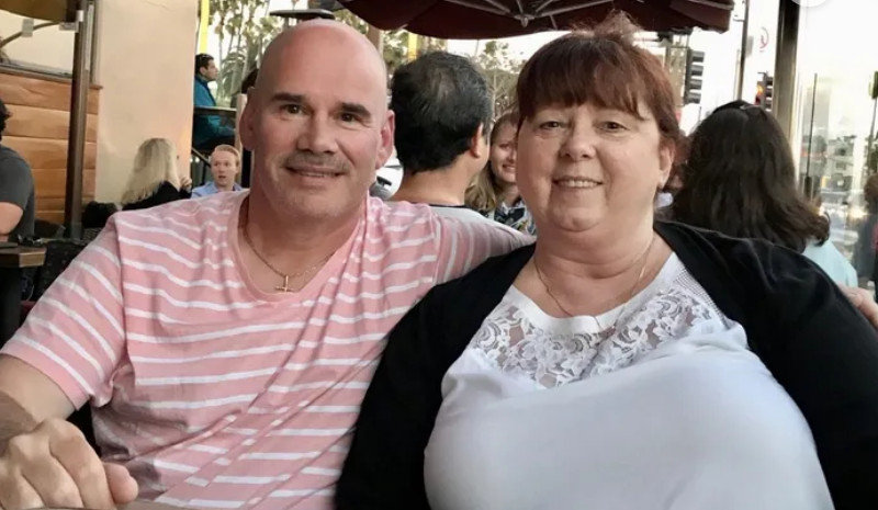 Bob Haines and his wife, Dawn, are seated at an outdoor establishment. Bob has light skin and a bald head and wears a light pink striped shirt. Dawn has light skin and red-brown hair pulled back and wears a white lace-trimmed shirt and a black sweater.