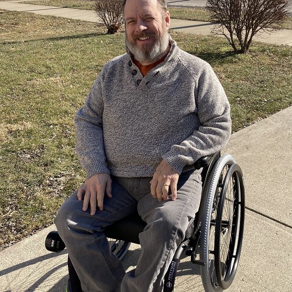 Living with hypophosphotasia, David Talkington is seated in a black manual chair wearing gray pants and a gray sweater. He is smiling with a salt and pepper beard and short hair.