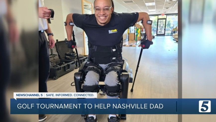 Eric Murray grins as he uses the ReWalk exoskeleton device 10 years after a spinal cord injury. The device is strapped to his legs with two black braces for his arms. Eric has light brown skin, glasses, and a bald head.