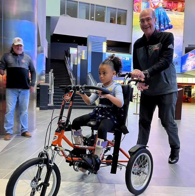 60-year-old Evangeline sits on her new black and orange adaptive bike and gets a push from a smiling Brad Marsh from the Flyers Alumni.