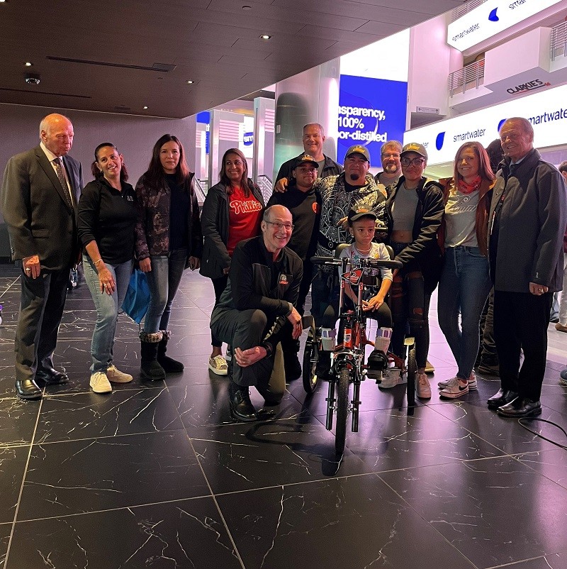 A group of family members, supporters, and Philadelphia Flyers alumni gather around 6-year-old Evangeline Torres on her new adaptive bike.