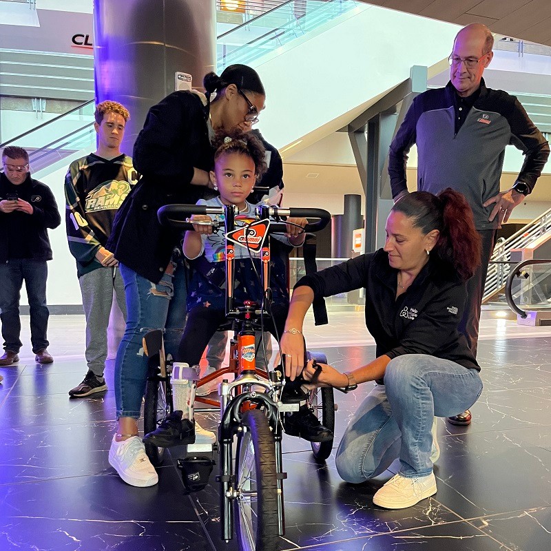 6-year-old Evangeline sits on her new black and orange adaptive bike as her mom, Kelly Green from Help Hope Live, and Brad Marsh from the Flyers Alumni help out.