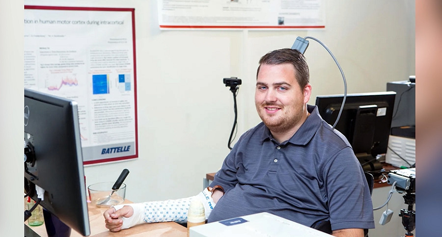 Ian Burkhart is seated at a desk with a brain-computer interface device that resembles a plug protruding from the back of his head and his right arm resting on the desk to interact with the device.