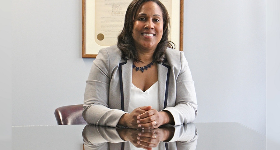 Keir sits at a desk with a reflective surface with a smile and her hands folded in front of her. She has brown skin and shoulder-length brown hair.