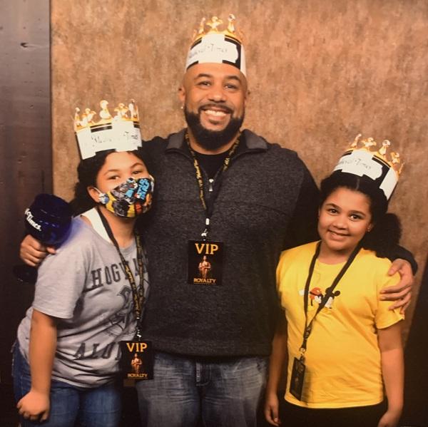 Transplant recipient Matthew poses with his two daughters wearing pretend crowns. Matthew has brown skin and a black short beard.