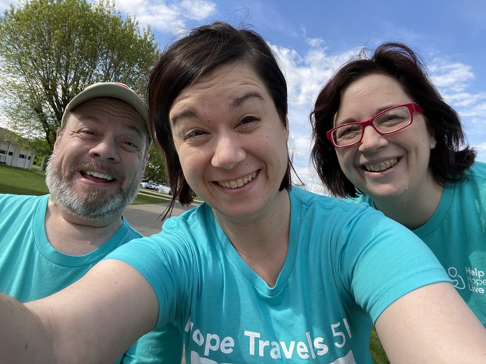 Sharon and David Talkington, Help Hope Live Ambassadors and a family facing hypophosphotasia, smile with their daughter between them wearing matching Hope Travels 5K light teal t-shirts.