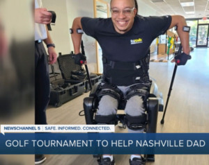 Eric Murray grins as he uses the ReWalk exoskeleton device 10 years after a spinal cord injury. The device is strapped to his legs with two black braces for his arms. Eric has light brown skin, glasses, and a bald head.