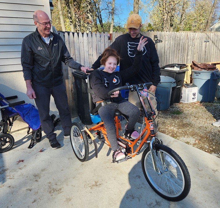 Flyers Alumni Brad Marsh holds the back of the black and orange Flyers themed adaptive bike as 9-year-old Raelynn sits on the bike and raises one arm into the air. Raelynn and a family member wear matching black Philadelphia Flyers jerseys. They are in a concrete driveway.