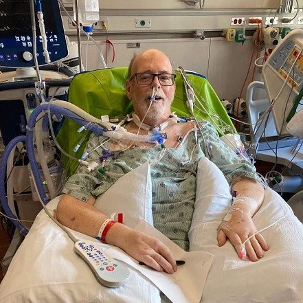 Double lung transplant recipient Carl Allison is pictured in the hospital post-transplant with a breathing tube hooked up to many wires and machines. He has light skin and a short white goatee with black-rimmed glasses.