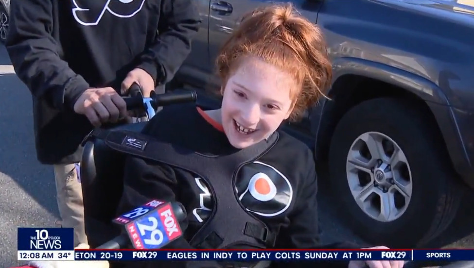 9-year-old Raelynn expresses her excitement about her new bike into a FOX 29 microphone held by a reporter off-screen. Raelynn has light skin and red hair and wears a black and orange Flyers jersey.