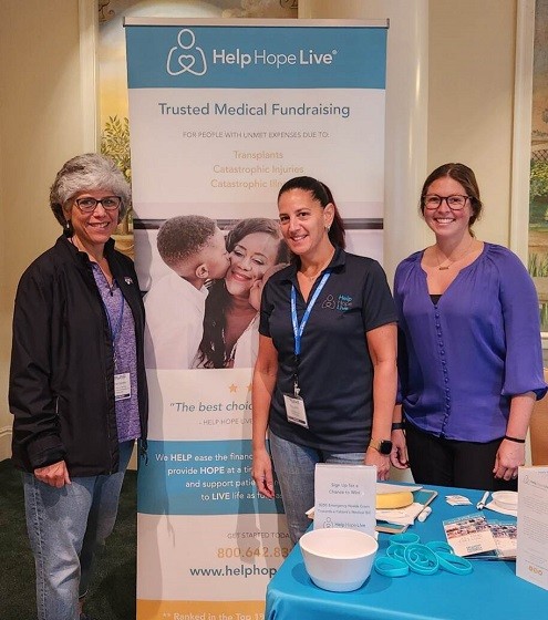 Kelly Green with a transplant social worker and Help Hope Live's Kate Lacouture with a Help Hope Live banner and exhibitor table covered in a teal tablecloth.