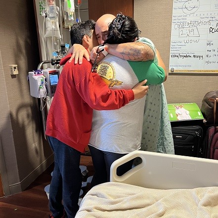 Heart translant recipient Kevin Lopez looks filled with emotion as he hugs his children pre-transplant. He has light brown skin and a bald head and is wearing a hospital gown.