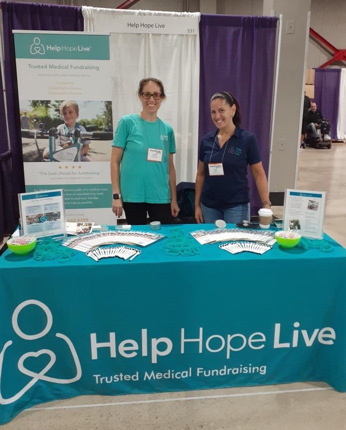 Kelly Green and Brooke Schostak stand at the Abilities Expo Miami 2022 booth for Help Hope Live behind a teal tablecloth and plenty of Help Hope Live brochures.