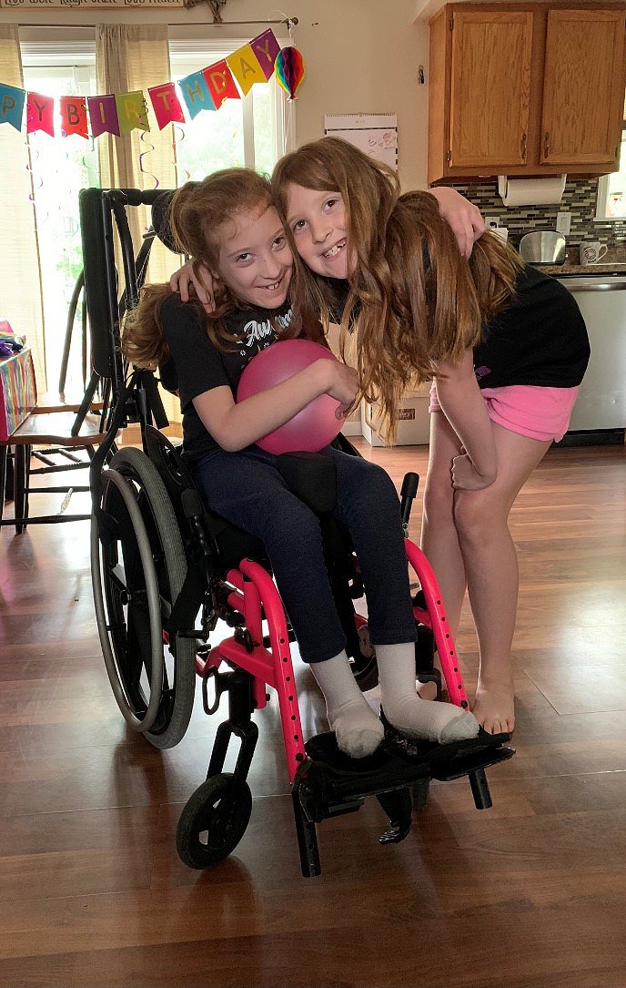 Raelynn, who is living with cerebral palsy, is pictured seated in her black and magenta wheelchair holding a pink ball on her lap. She is smiling with her arm around her sister. Raelynn and her sister both have light skin and long red hair with brown eyes.