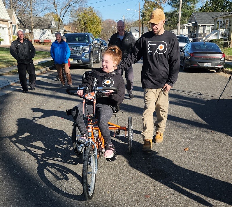 9-year-old Raelynn rides her new adaptive bike in the street outside of her home as Philadelphia Flyers Alumni Bob Kelly, Brad Marsh, and Joe Watson look on smiling. Raelynn is being pushed by a family member who wears a black Flyers jersey that matches hers.