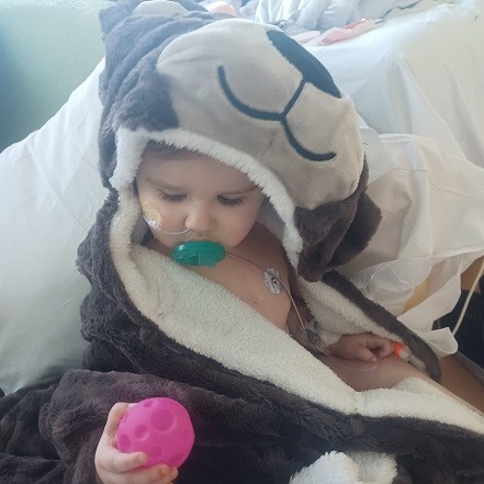 Liver transplant recipient River Cole is a 1-year-old with light skin and a teal pacifier. He is pictured post-transplant in a cozy hooded bear blanket with a tube in his nose.