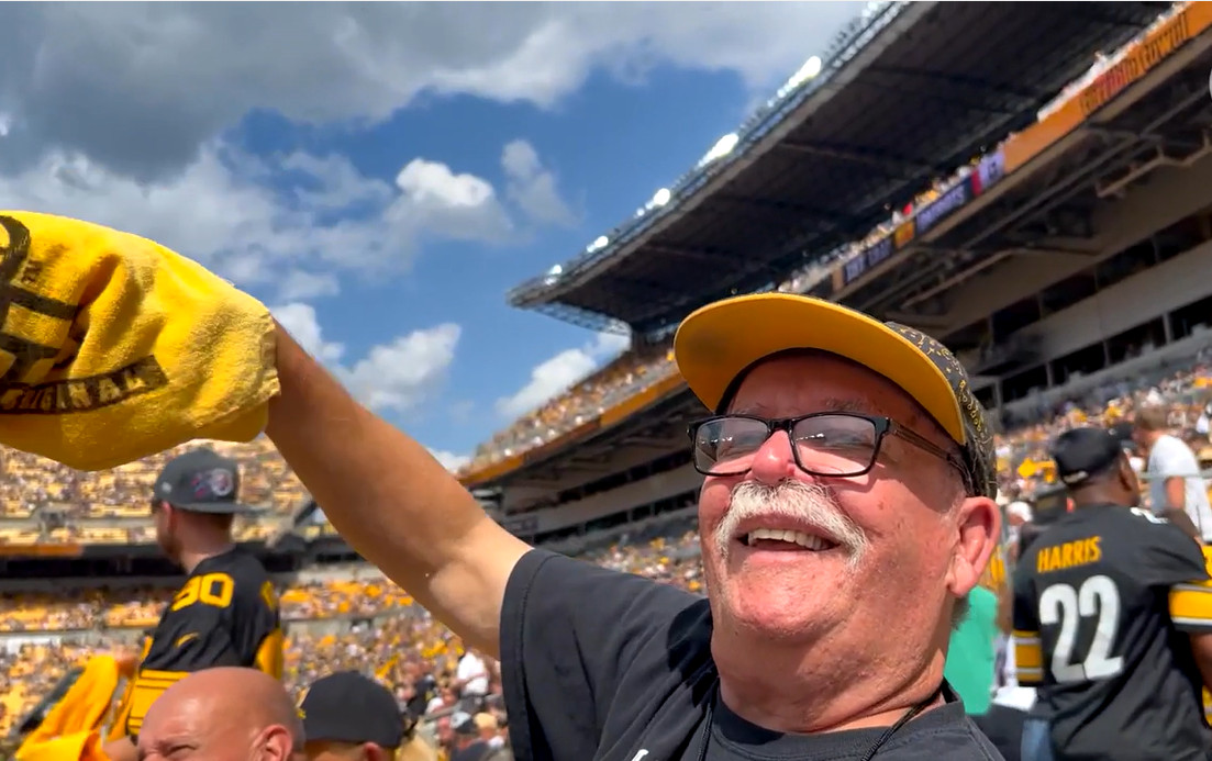 Zim Zimmerman smiles deeply as he waves a yellow Steelers towel at his first Steelers game. He has light skin, a white mustache, black-rimmed glasses, and a black shirt with a Steelers ball cap.