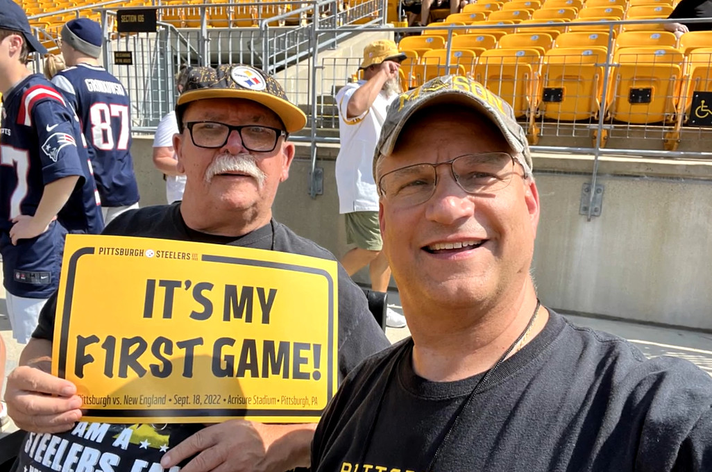 Jim Zimmerman wears a Steelers hat and holds a bright yellow sign that reads it's my first game! Beside him is heart transplant recipient Bill Soloway in a black t-shirt with a ball cap.