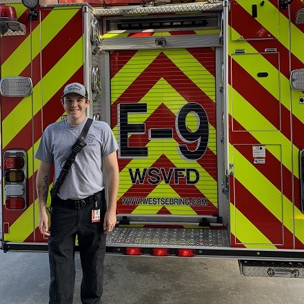 Kidney transplant recipient Zachary Campbell is pictured in his volunteer uniform with a gray shirt and black pants in front of an emergency vehicle that reads E-9 WSVFD www.westsebring.com