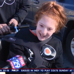 9-year-old Raelynn expresses her excitement about her new bike into a FOX 29 microphone held by a reporter off-screen. Raelynn has light skin and red hair and wears a black and orange Flyers jersey.