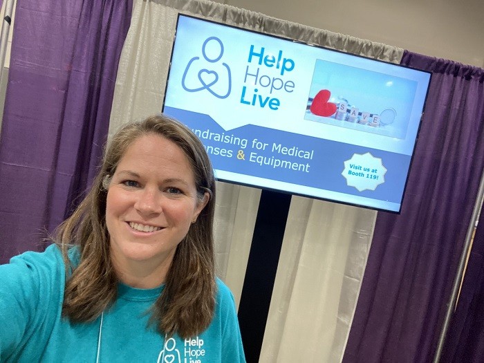 Sonny wears a teal Help Hope Live t-shirt as she stands in front of a TV screen displaying a promotion for a Help Hope Live medical fundraising workshop at a conference. She has light skin and sandy brown shoulder-length hair.