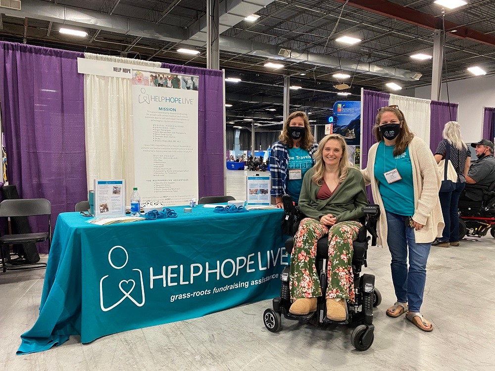 Sonny and Kate wear teal Help Hope Live shirts and black face masks as they stand in front of the Help Hope Live booth at a conference with client and ambassador Lauren Shevchek, who is seated in her black power chair with light skin and blonde hair just past her shoulders.