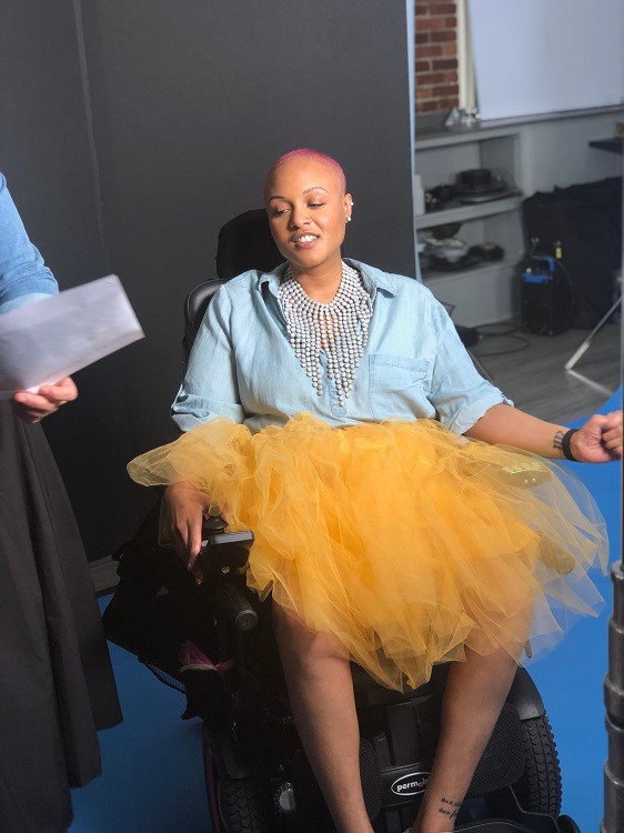 Keisha Greaves looks down in a photoshoot setting with a blue backdrop behind her. She is seated in her black power wheelchair with a yellow tulle tutu and a blue soft denim-like top with a dramatic pearl-like statement necklace. She has light brown skin, a stud in her nose, and very short pink dyed hair.