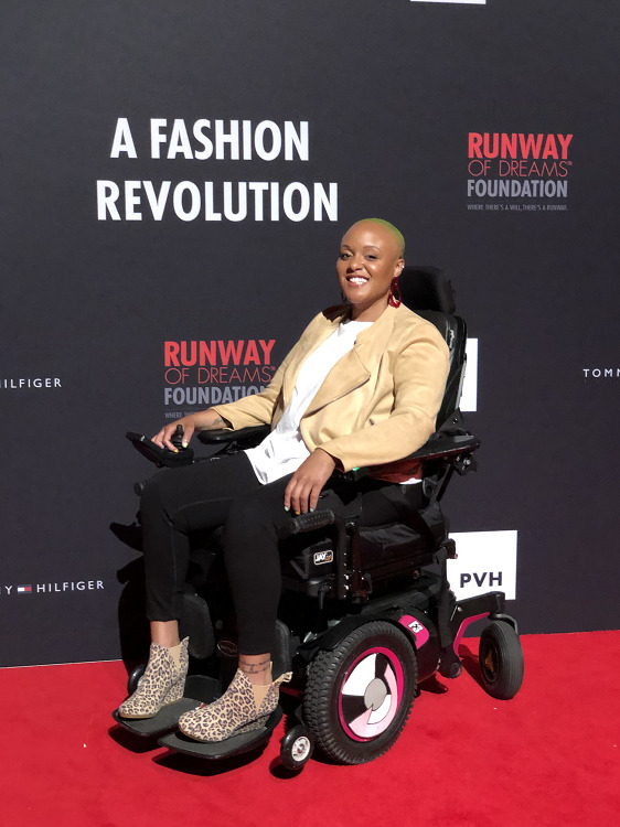 Keisha Greaves is on a red carpet with a backdrop that reads Runway of Dreams Foundation A Fashion Revolution. Seated in her black power wheelcahir, she wears leopard print booties, black pants, a white shirt, and a leather-like light brown jacket. She has light brown skin, dark eyes, red dangling earrings, and very short green dyed hair.