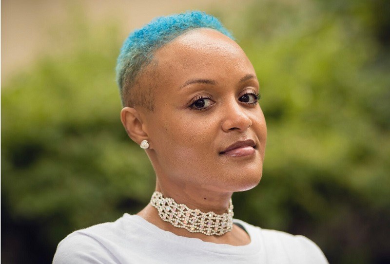 In a horizontal portrait, Keisha Greaves looks into the camera with dark brown eyes, light brown skin, short curly blue hair, and sparkling jewelry at her ears and throat.