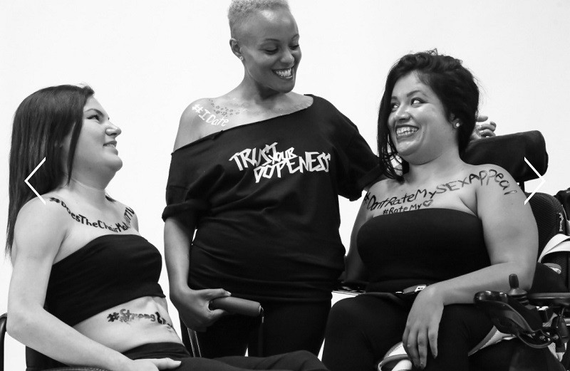 In a black and white photo, Keisha Greaves stands with her mobility aid in her right hand. She has light brown skin and short curly hair with a black off-the-shoulder shirt from her Girls Chronically Rock collection that reads Trust Your Dopeness. Two women to her right and left smile at her and wear black sleeveless tops. All three women have hashtags painted on her body: visible are #IDate and DontRateMySexAppeal #RateMyHeart