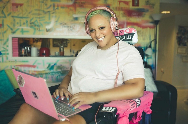 Keisha Greaves is seated in her black power wheelchair with a pink laptop on her lap and metallic pink headphones. She wears a white shirt and has light brown skin, a stud in her nose, dark brown eyes, and short curly blue and green dyed hair.