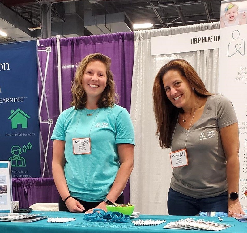 Kate Lacouture and Kelly Green from Help Hope Live wear Abilities Expo nametags and smile as they stand behind a Help Hope Live teal table.