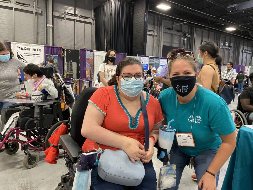 Sonny Mullen leans down so she is the same height as activist Emily Ladau, who wears a bright orange shirt and is seated in her black power chair. Both women are wearing face masks as they attend an Abilities Expo event.