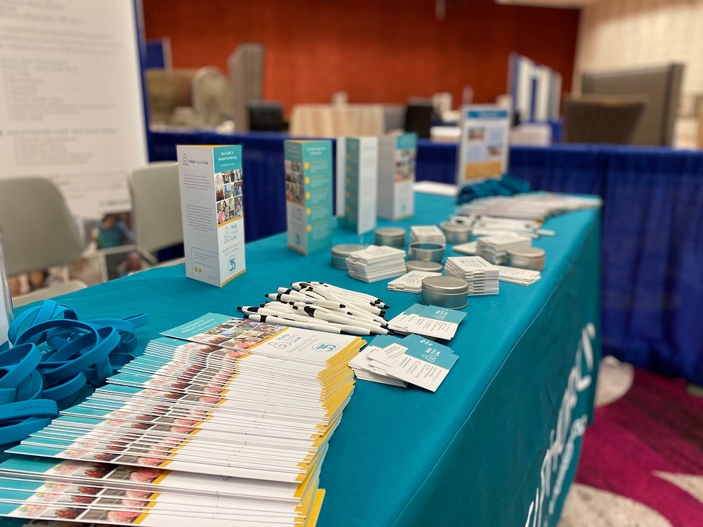 A conference table for Help Hope Live displays a teal tablecloth and large stacks of brochures, business cards, pens, and teal bracelets.